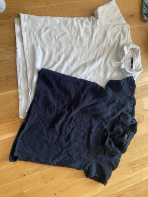 Två pikétröjor i svart och beige - Säljer två pikétröjor i ribbad bomull, en i svart och en i beige. Båda har korta ärmar, klassisk krage och knappar vid halsen. Perfekta för en avslappnad och stilren look.