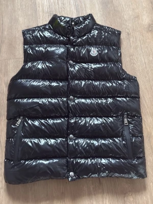 Svart glansig dunväst Moncler - Snygg svart dunväst från Moncler med glansigt material och hög krage. Västen har två dragkedjeförsedda fickor framtill och tryckknappar. Klassisk Moncler-logga på bröstet. Perfekt för lager-på-lager och streetstyle.