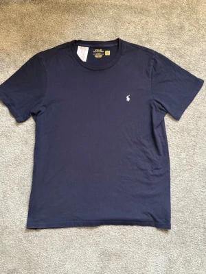 Mörkblå t-shirt från Polo Ralph Lauren - NÄSTAN HELT NY, ANVÄND MAX 5 GÅNGER! Klassisk mörkblå t-shirt från Polo Ralph Lauren med rund halsringning och den ikoniska broderade loggan på bröstet. Tillverkad i mjuk bomull och har en normal passform. Perfekt för en stilren och avslappnad look.