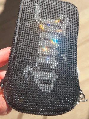 Mobilväska / aftonväska i strass. Juicy Couture - Helt ny & oanvänd svart axelväska / aftonväska / mobilväska från Juicy Couture med glittriga stenar och stor Juicy-logga i silver på framsidan.  Väskan har en silverfärgad kedja som axelrem och en snygg dragkedja med Juicy-detalj.  •Min iPhone 14 Pro med skal fick perfekt plats i väskan•                                               Cirka mått i cm: Höjd: 16-17 Bredd: 10 ( den bredaste sidan) Djup: 3. 
