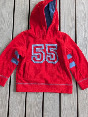 Röd hoodie med blått nummer 55, barn stl100 - Snygg röd hoodie i fleece med stor huva och blåa detaljer. På bröstet finns ett stort, broderat nummer 55 i blått och vitt. Ärmarna har blåa ränder som ger en sportig look. Perfekt för dig som gillar streetwear och vill sticka ut.
