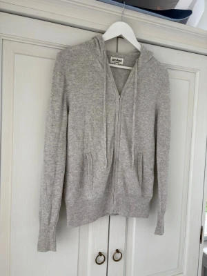 Ljusgrå kofta i kashmir från Soft Goat - Mysig ljusgrå zip up i kashmir från Soft Goat. Dragkedja framtill, två fickor och  huva med snören. Perfekt för dig som gillar stilrena och bekväma plagg. Ribbstickade muddar vid ärmslut och nederkant.