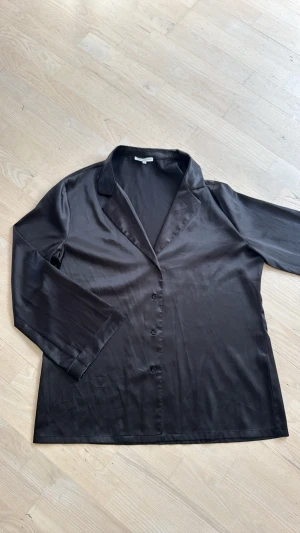 Sanne Alexandra Svart skjorta i satin-look - Stilren svart skjorta med lång ärm och knappar framtill. Skjortan har en slät, glansig satin-liknande yta och klassisk krage. Perfekt för dig som gillar en clean och elegant vibe.