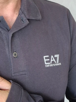 Mörkgrå långärmad skjorta EA7 storlek M  - Snygg mörkgrå långärmad skjorta från EA7 Emporio Armani med klassisk krage och knappar framtill. Skjortan har en stilren logga på bröstet och är tillverkad i mjukt bomullsmaterial. Perfekt för en clean och avslappnad look. Färgen har bleknat något på grund av användning, men den behåller fortfarande sin elegans.