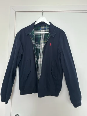 Marinblå bomberjacka från Polo Ralph Lauren - Klassisk marinblå bomberjacka från Polo Ralph Lauren med röd logga på bröstet. Jackan har dragkedja framtill, två snedställda fickor och rutigt innerfoder i grönt och vitt. Tillverkad i bomull och har långärmad design.