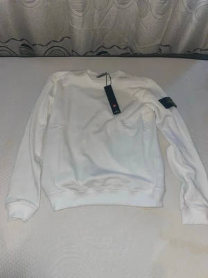 Vit sweatshirt från Stone Island - Vit sweatshirt från Stone Island med klassisk rund hals och ribbade muddar. Tröjan har den ikoniska logotypen på vänster ärm och är tillverkad i mjukt material för en clean och stilren look.