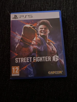 Street Fighter 6 till PS5 - Street Fighter 6 till PlayStation 5 i blå plastfodral. Omslaget har en färgstark illustration med en karaktär i fightingpose och spelets logga. Rekommenderad ålder är 12 år. Perfekt för dig som gillar actionfyllda fightingspel.