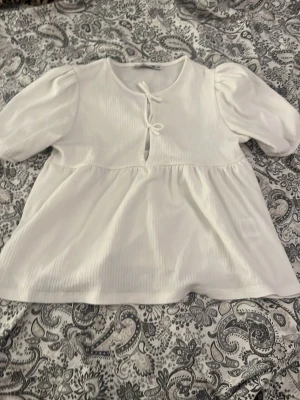 Vit blus med puffärm och knyt - Söt vit blus med puffiga ärmar och knyt framtill vid halsen. Blusen har en lätt rynkad midja och ett luftigt fall. Materialet är tunt och har en diskret struktur som ger en extra touch. Perfekt till jeans eller kjol för en trendig look.💗
