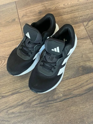 Svarta Adidas sneakers med vita detaljer - Snygga svarta sneakers från Adidas med klassiska vita ränder på sidan och vit sula. Ovandelen är i meshmaterial för bra andningsförmåga och skön komfort. Perfekta för dig som vill ha en sportig och stilren look.