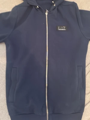 Marinblå hoodie med dragkedja EA7 - Snygg marinblå hoodie från EA7 med dragkedja framtill och två fickor. Hoodien har en huva med dragsko och en diskret EA7-logga på bröstet. Perfekt för en avslappnad och sportig stil.