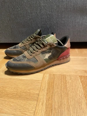 Valentino Rockrunners  - Snygga sneakers från Valentino Garavani med camomönster i grönt, beige och svart. Skorna har detaljer i mocka och läder samt en röd hälkappa med nitar. Perfekta för dig som vill sticka ut med en unik och trendig look.