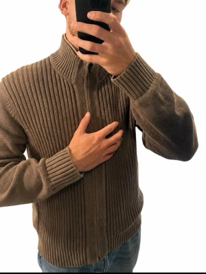 Tommy Hilfiger cardigan str L  - Stickad ziptröja med hög krage – storlek L  Stilren och kraftig stickad tröja i en snygg brungrå/taupe nyans. Hel dragkedja, hög ribbad krage och ribbstickade detaljer framtill ger en exklusiv och välklädd look.  Varm och bekväm – perfekt till höst och vinter. Enkel att kombinera med både jeans och finare byxor.  Storlek L. Modellen är 186 cm – se passformen på bilden.  #stickadtröja #ziptröja #herrtröja #storlekL #höstmode #vintermode #herrmode #knitwear #zipcardigan #taupe #bruntröja #minimali