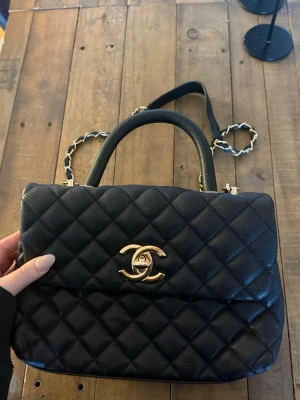 Svart Chanel quiltad handväska - Säljer en svart quiltad handväska från Chanel med klassiskt guldigt CC-spänne framtill. Väskan har både topphandtag och axelrem i kedja med läderdetaljer. Tillverkad i läder med elegant rutmönster. Perfekt för dig som vill ha en ikonisk och lyxig accessoar.