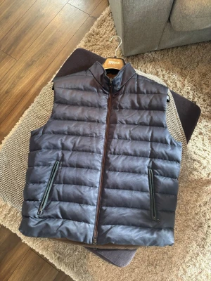 Loro Piana Väst  - Snygg mörkblå dunväst ifrån Loro Piana med quiltad design och dragkedja framtill. Västen har två praktiska fickor med dragkedja och hög krage. Insidan är brun med orange detaljer och innerfickor. Perfekt för lager-på-lager under kyliga dagar. Som ny, inget slitage, tillkommer box