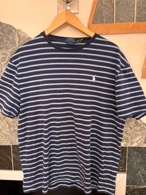 Randig t-shirt från Polo Ralph Lauren - Mörkblå t-shirt med ljusblå horisontella ränder från Polo Ralph Lauren. Klassisk passform med korta ärmar och rund halsringning. Ikoniska loggan broderad på bröstet. Perfekt till jeans eller shorts.