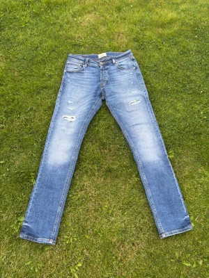 Jack & Jones jeans  - Säljer ett par blå skinny jeans med slitningar och ljusa tvättade partier. Jeansen har klassisk femficksmodell, låg midja och dragkedja med knapp. Materialet är denim i bomull. Perfekta för en avslappnad och trendig look.