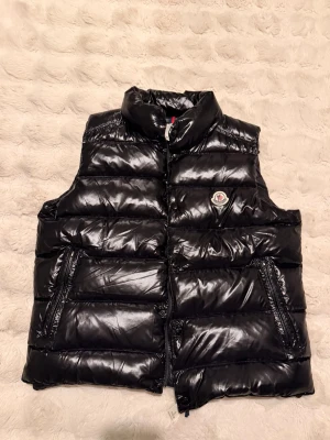 Svart dunväst från Moncler - Snygg svart dunväst från Moncler med glansig finish och hög krage. Västen har två dragkedjeförsedda fickor framtill och ett broderat Moncler-märke på bröstet. Perfekt för lager-på-lager och ger en trendig look.