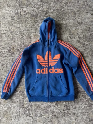 Blå Adidas hoodie med orange tryck - Snygg blå hoodie från Adidas med dragkedja, stor orange logga och text på bröstet samt klassiska ränder längs ärmarna. Utrustad med huva och två fickor framtill. Perfekt för en sportig och avslappnad stil.