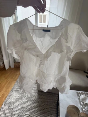 Vit volangblus från Zara - Säljer en superfin vit blus från Zara med korta ärmar och volangdetaljer längs urringningen och fållen. Blusen har en luftig, skrynklig struktur och djup V-ringning. Perfekt för en trendig och avslappnad look.🤍
