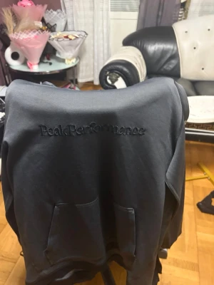 Mörkgrå hoodie från Peak Performance - Snygg mörkgrå hoodie från Peak Performance med broderad logga på bröstet. Klassisk känguruficka framtill och huva. Perfekt för en avslappnad och sportig stil. Tillverkad i mjukt material som känns skönt mot huden.