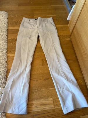 Beiga kostymbyxor med raka ben - Snygga beiga kostymbyxor med raka ben och pressveck. Byxorna har diskreta fickor fram och hällor för skärp i midjan. Perfekta för en clean och stilren look. Materialet ser ut att vara en mjuk och lätt blandning, vilket ger en skön känsla.