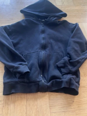 Svart hoodie med dragkedja från H&M - Klassisk svart hoodie från H&M med dragkedja framtill och stor huva. Tröjan har ribbade muddar vid ärmslut och nederkant samt en stor magficka. Perfekt för en avslappnad och enkel stil.