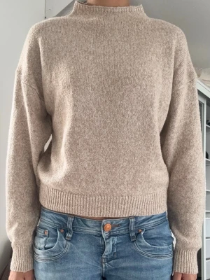 Beige stickad tröja med hög hals - En enkel och stilren beige stickad tröja med hög hals och långa ärmar. Tröjan har ribbade muddar vid ärmslut och nederkant. Perfekt för lager på lager och passar till det mesta i garderoben.