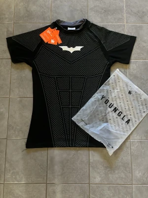 Svart Batman träningströja YoungLA - Svart comp shirt från YoungLA med Batman-logga på bröstet och ett detaljerat mönster som liknar en superhjältedräkt. Sitter bra för dig som har M