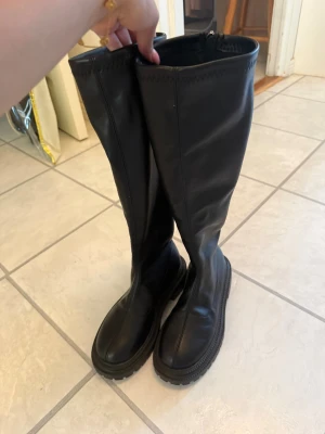 Svarta höga boots i skinn - Säljer ett par svarta höga boots i slätt skinn med grov sula och dragkedja baktill. Stövlarna har en stilren design och passar perfekt till både jeans och kjol. Klassisk modell som är enkel att matcha och ger en edgy look.