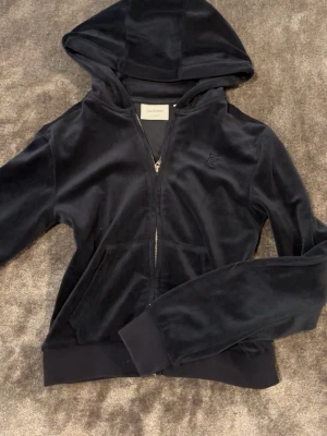 Svart velour hoodie med dragkedja - Mjuk och skön svart hoodie i velour med dragkedja framtill och fickor på sidorna. Har en huva och ribbade muddar vid ärmslut och nederkant. Perfekt för en avslappnad och trendig look.