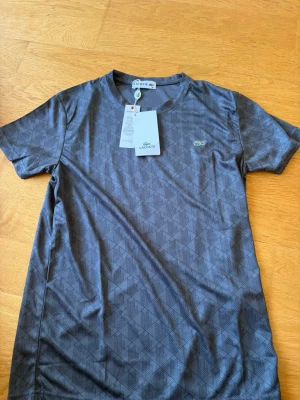 Grå mönstrad t-shirt från Lacoste - Snygg grå t-shirt från Lacoste med diskret geometriskt mönster och klassisk rund hals. T-shirten har korta ärmar och den ikoniska gröna krokodilloggan på bröstet. Perfekt till jeans eller shorts för en clean och stilren look.