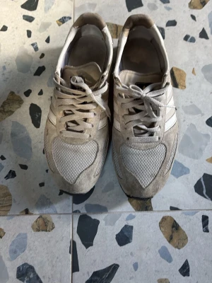Beige Adidas sneakers med meshdetaljer - Säljer ett par beige Adidas sneakers med klassiska vita ränder på sidan. Skorna har ovandel i mocka och mesh för en sportig look och skön känsla. Yttersulan har gummidobbar för extra grepp. Perfekta för dig som gillar retrostil och streetwear.