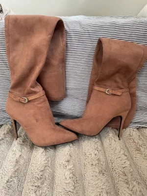   Overknee mocka stövlar   - Snygga brun rosa  overknee stövlar i mockaimitation med spetsig tå och smal hög klack. Stövlarna har ett dekorativt spänne runt ankeln och är perfekta för att lyfta vilken outfit som helst. Materialet är mjukt och följsamt, vilket ger en skön passform.