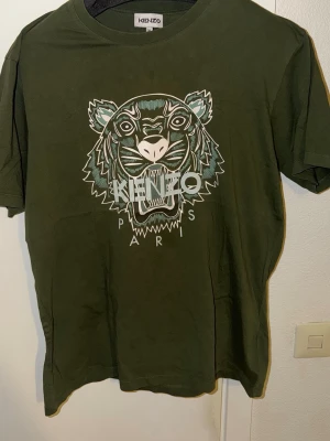 Grön Kenzo t-shirt med tigertryck - Snygg grön t-shirt från Kenzo med det ikoniska tigertrycket och texten 'Kenzo Paris' på bröstet. T-shirten har rund hals och korta ärmar. Tillverkad i mjuk bomull som känns skön mot huden. Perfekt för dig som vill sticka ut med en cool design.