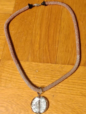 Brunt pärlhalsband med silverfärgat hänge - Snyggt halsband med små bruna pärlor i ett tätt mönster och ett runt, silverfärgat hänge med unik struktur. Halsbandet har ett diskret metallspänne och känns både trendigt och personligt. Perfekt för dig som gillar detaljer och vill sticka ut lite extra.