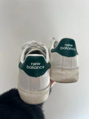 Vita New Balance sneakers med gröna detaljer - Snygga vita sneakers från New Balance med gröna detaljer på hälen och klassisk logga. Skorna har lågt skaft och är tillverkade i textil och syntetmaterial. Perfekta för dig som gillar en clean och sportig stil.