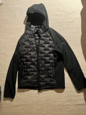 Svart lättsviktsjacka med huva - Snygg svart pufferjacka med huva och dragkedja framtill. Jackan har quiltad front och släta ärmar samt sidor, vilket ger en modern look. Perfekt för kalla dagar när du vill hålla stilen enkel och clean.