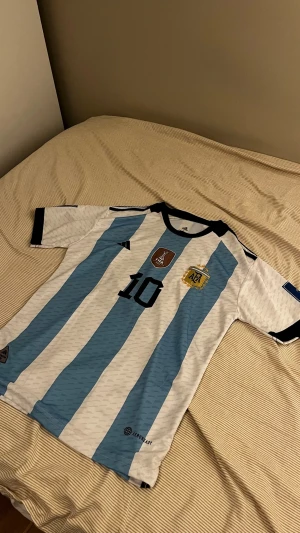 Argentina Messi matchtröja blåvit - Säljer en Argentina matchtröja med Messi och nummer 10. Tröjan har klassiska blå och vita ränder, svarta detaljer vid krage och ärmslut samt officiella tryck och emblem på bröstet. Perfekt för dig som älskar fotboll och vill stötta landslaget.