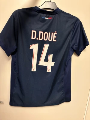 PSG matchtröja D. Doué #14 - Snygg mörkblå PSG matchtröja från Nike med D. Doué och nummer 14 på ryggen. Tröjan har röd och vit rand på framsidan, PSG-logga och Qatar Airways sponsortryck. Perfekt för fotbollsfans som vill sticka ut på läktaren eller planen.