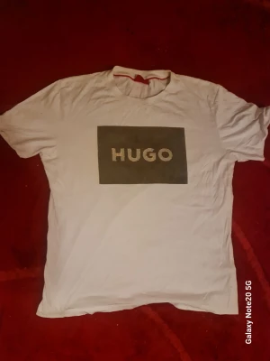 Vit t-shirt från HUGO med logga - Vit t-shirt från HUGO med stor svart logga på bröstet. Klassisk rund halsringning och korta ärmar. Tillverkad i mjukt bomullsmaterial för en bekväm känsla. Perfekt för en clean och stilren look.