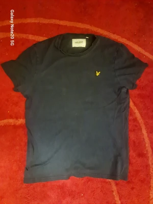 Mörkblå t-shirt från Lyle & Scott - Snygg mörkblå t-shirt från Lyle & Scott med klassisk passform och korta ärmar. Tillverkad i ekologisk bomull och prydd med den ikoniska gula loggan på bröstet. Perfekt för en stilren och avslappnad look.