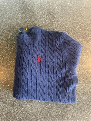 Mörkblå kabelstickad tröja  - Kabelstickad mörkblå tröja med rund halsringning och röd broderad logga på bröstet. Tröjan sitter som en xs i storleken