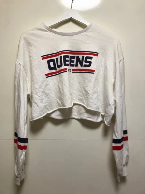Vit croppad långärmad topp QUEENS - Vit croppad långärmad topp med trycket 'QUEENS NYC' framtill. Röd och marinblå rand på bröstet och ärmarna. Mjuk bomullskänsla och relaxed fit, perfekt till jeans eller kjol för en sportig streetstyle.