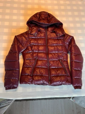 Vinröd Lager 157 jacka  - Snygg vinröd pufferjacka med glansig finish och avtagbar huva. Jackan har två snedställda fickor med dragkedja och quiltad design. Perfekt för kalla vinterdagar och ger en trendig look.