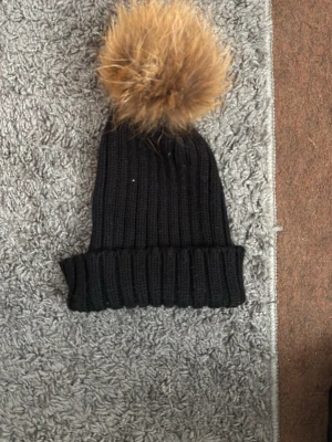 Svart ribbstickad mössa med pälsboll - Snygg svart ribbstickad mössa med bred uppvikt kant och stor fluffig pälsboll i brunt. Perfekt för kalla dagar när du vill hålla stilen. Mössan är gjord i mjukt, stretchigt material och har en modern look. 20 kronor styck för en och för alla så är priset 100kr