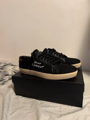 Svarta Saint Laurent sneakers - Säljer ett par svarta Saint Laurent sneakers med vit text på sidan och beige sula. Skorna har klassisk låg profil, svarta skosnören och är tillverkade i en mix av läder och textil. Perfekta för dig som gillar stilrena och exklusiva sneakers.