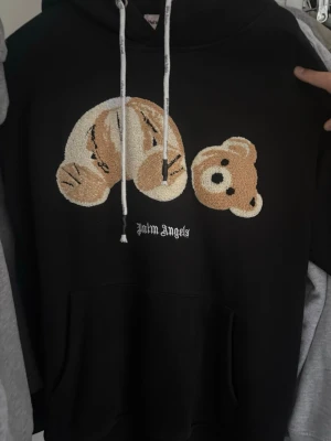 Svart hoodie Palm Angels med björn - Svart hoodie från Palm Angels med en stor broderad björn på bröstet och vit text. Hoodien har huva med vita snören och en klassisk känguruficka framtill. Materialet är mjukt och skönt, perfekt för en avslappnad streetwear-look.