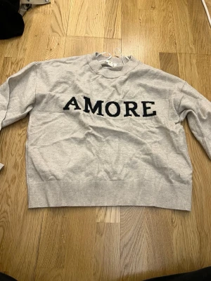 Beige sweatshirt med svart text AMORE - Beige sweatshirt från Gina Tricot med svart texten 'AMORE' framtill. Tröjan har rund halsringning, ribbade muddar och avslappnad passform. Perfekt för en chill och trendig look.