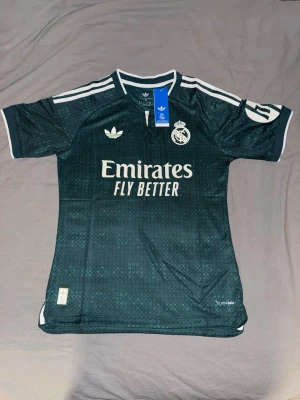 Mörkgrön Real Madrid tröja Adidas - Snygg mörkgrön Real Madrid matchtröja från Adidas med vita detaljer och klubbmärke på bröstet. Tröjan har korta ärmar, rund hals och är tillverkad i ett lätt, ventilerande funktionsmaterial. Perfekt för fotbollsträning eller som supporterplagg.