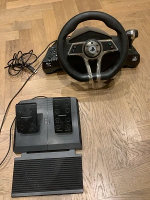 Ratt och pedaler till PlayStation - Säljer en gamingratt med pedaler för PlayStation. Ratten har svart grepp i konstläder och metallinslag, samt knappar för styrning. Pedalerna är svarta i plast med gummiyta för bättre grepp. Perfekt för racing- och bilspel för en mer realistisk spelupplevelse.